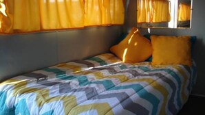 A single bed with a striped bedspread and two pillows, a window with yellow curtains, and a mirror on the wall. - "Lucille" Vintage Glamper Hideaway (Joshua Tree)