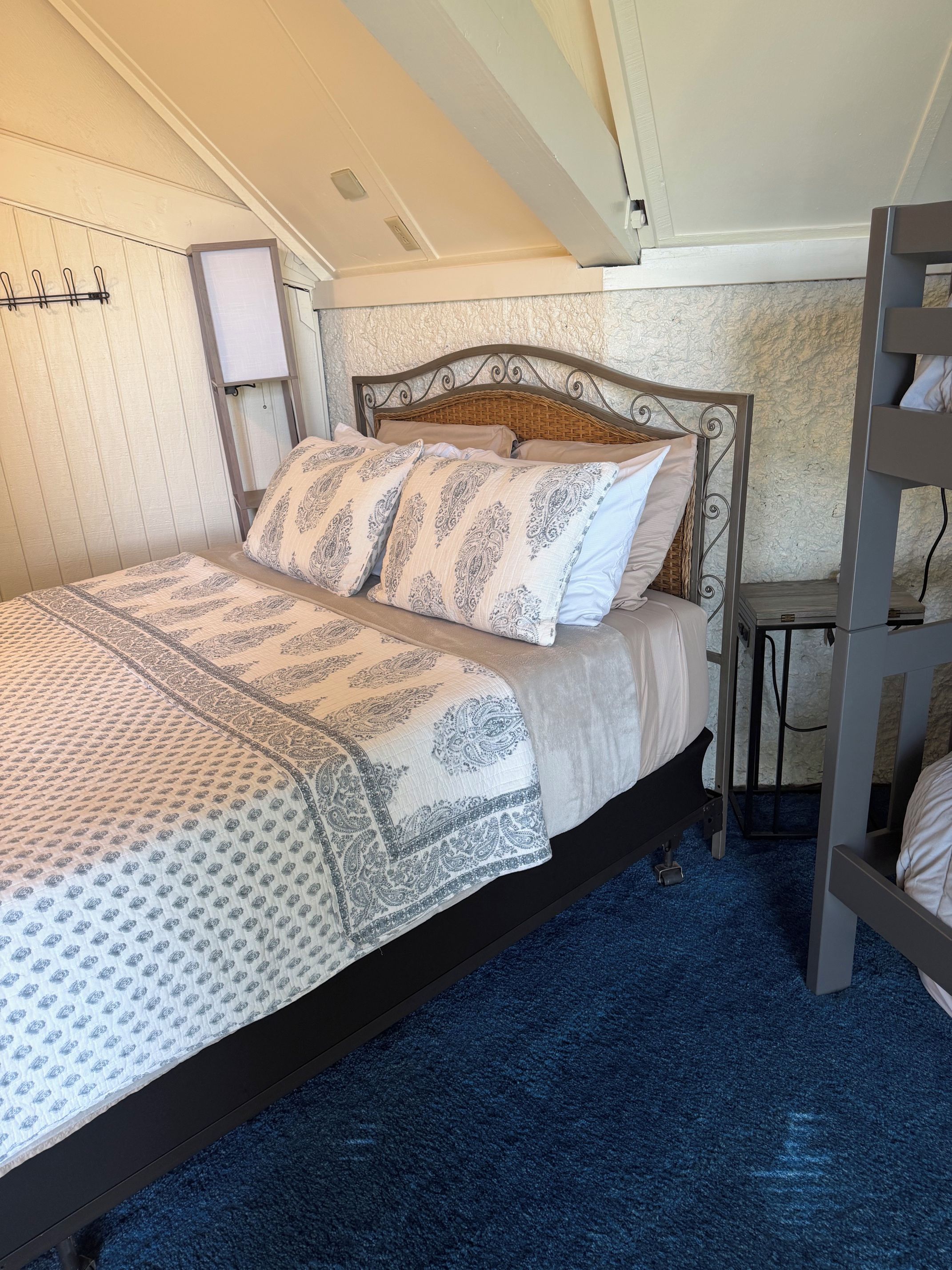 Een eenpersoonsbed met een bedspread met patroon en kussens, een nachtkastje, een lamp en een stapelbedconstructie.