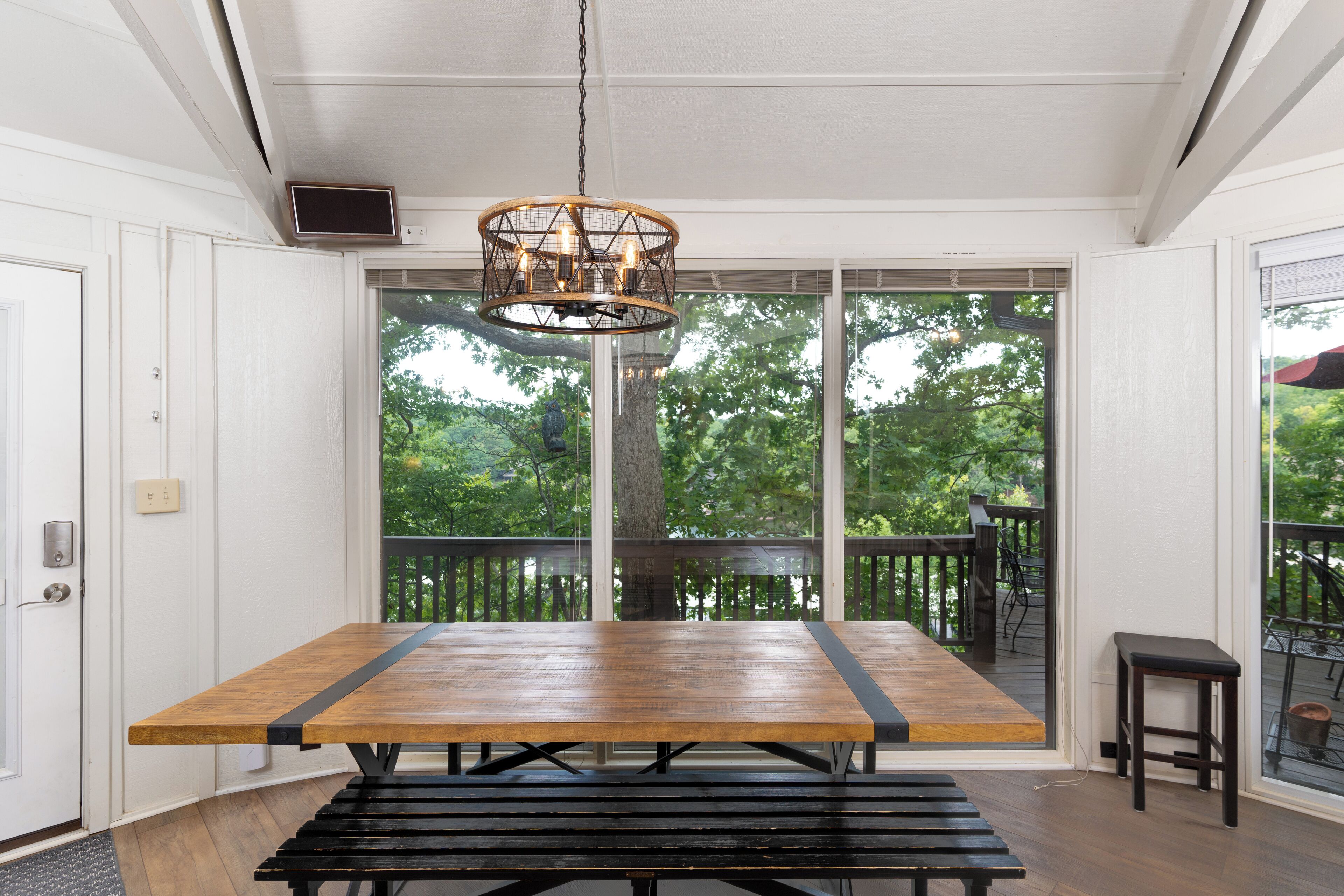 A dining area with a wooden table and bench, a hanging light fixture, and a view of a deck and trees through large glass doors.