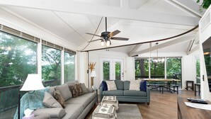 A spacious living room with a gray sofa, a blue couch, and a wooden coffee table. There is a ceiling fan, large windows, and a view of trees outside.
