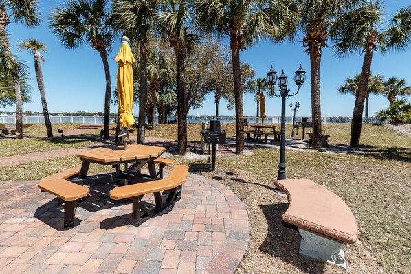 A picnic area with tables and benches, surrounded by palm trees and a view of the water. - Sunny Corner Condo, Ground Level, Direct Lazy River Access, Free Beach Service! (Fort Walton Beach)