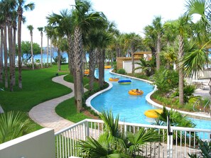 A pool area with a winding water feature, surrounded by palm trees and a view of the ocean. - Sunny Corner Condo, Ground Level, Direct Lazy River Access, Free Beach Service! (Fort Walton Beach)