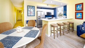 Een lichte keuken met witte kasten, een magnetron en een koelkast. Er staan vier rieten stoelen bij een ronde tafel met blauwe placemats.