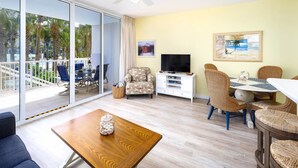 Een ruime woonkamer met een grote schuifpui naar een balkon, een houten salontafel, rieten meubels, een flatscreen-tv en kunstwerken aan de muur.