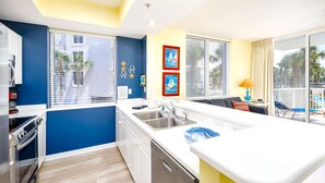 Een moderne keuken met een vaatwasser, oven en gootsteen. Twee ramen met zonwering, een balkon met uitzicht op palmbomen en een eethoek met tafel en stoelen.