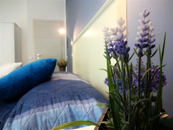 A bedroom with a bed, a blue pillow, and a potted plant with purple flowers. - I vicini della cattedrale (Palermo)