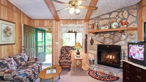A cozy living room with a fireplace, a television, a patterned sofa, a wooden ceiling fan, and a stone wall.