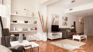 Un salon moderne avec un canapé, une table basse et un meuble pour la télévision. On y trouve des étagères ornées d’objets décoratifs et une grande fenêtre qui laisse entrer la lumière naturelle.