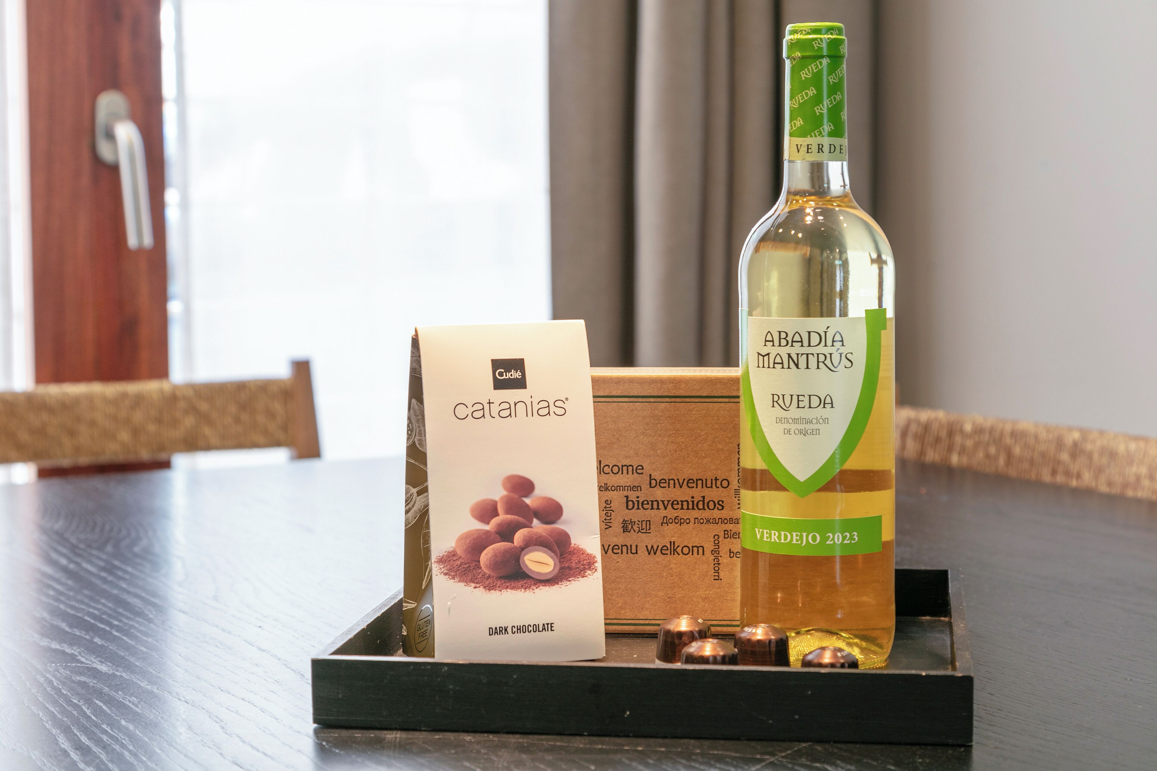 Une bouteille de vin Abadia Mantrús Verdejo 2023, une boîte de chocolats noirs sans gluten et un ensemble de cuillères à dégustation en cuivre, le tout présenté sur un plateau.