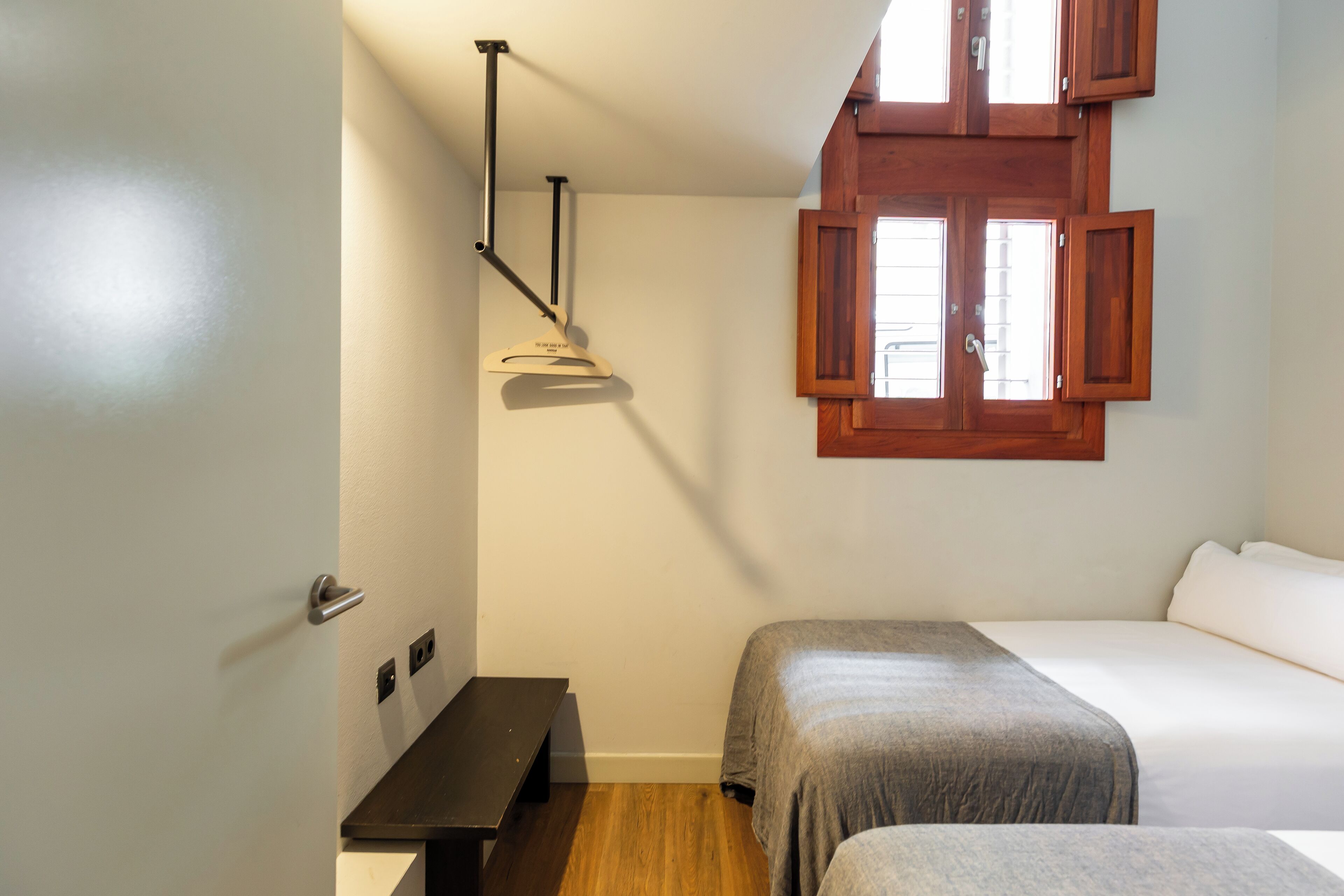 A bedroom with two beds, a wooden bench, a desk, and a window with wooden shutters.