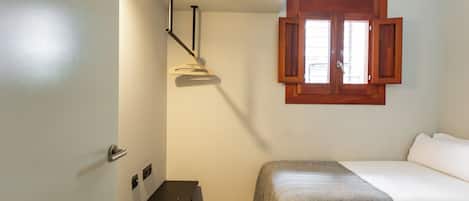 A bedroom with two beds, a wooden bench, a desk, and a window with wooden shutters.