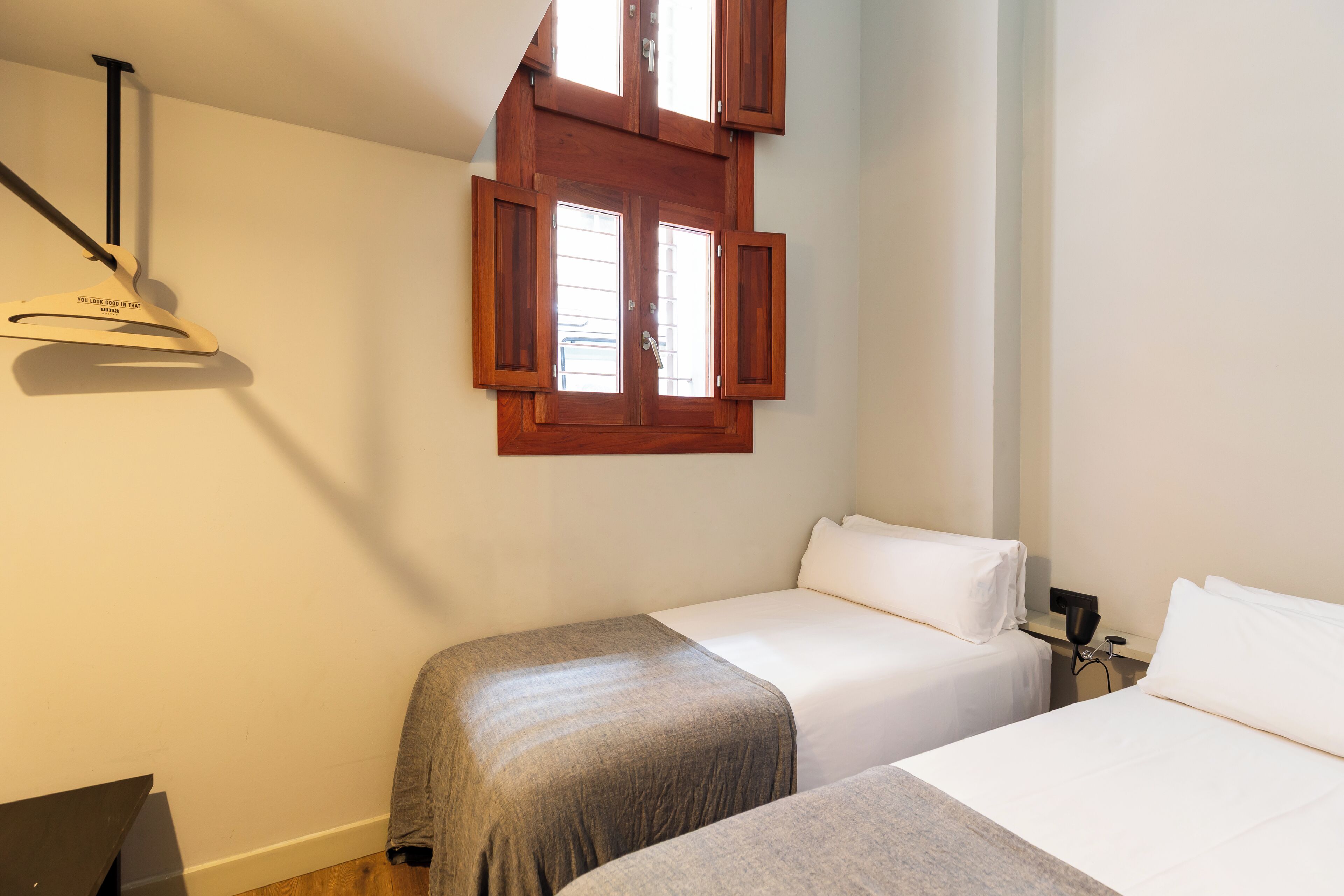 A hotel room with two beds, a wooden window with shutters, a desk with a lamp, and a ceiling-mounted light fixture.