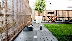 Un patio con piso de madera, una mesa y sillas blancas, y una planta grande en maceta.