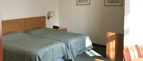 A hotel room with two beds, a wooden headboard, a small bedside table with a lamp, two framed pictures on the wall, and a chair with a table.