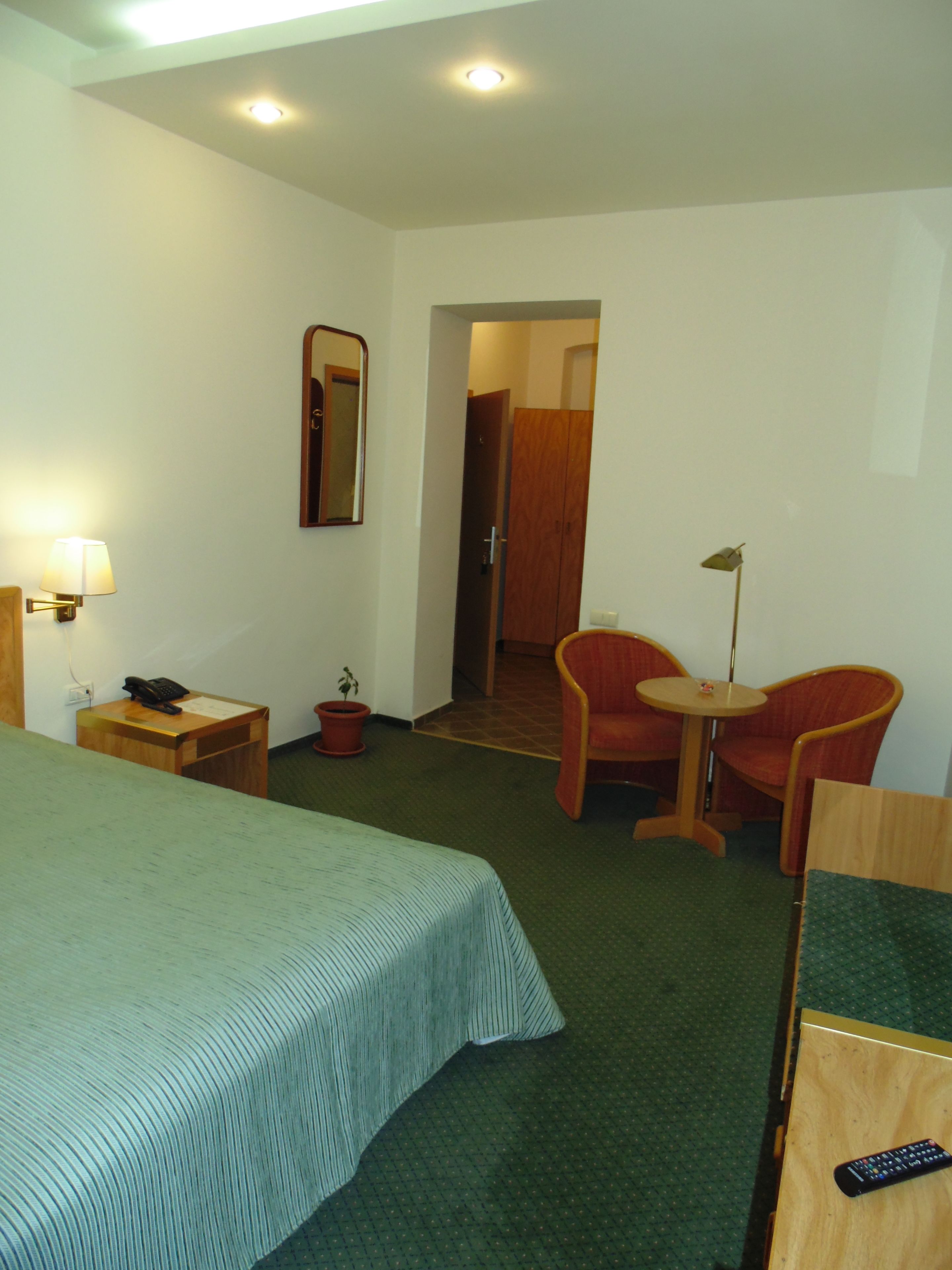 A hotel room with a bed, a desk with a lamp, a chair, a small table, and a mirror on the wall.