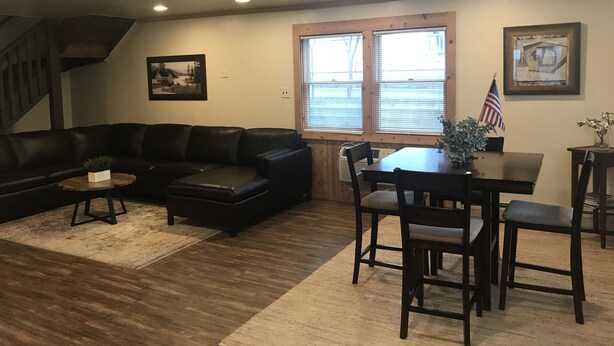 A living room with a black leather couch, a wooden dining table with chairs, a small coffee table, and a window with blinds. - The Lake Geneva Lakehouse (Williams Bay)