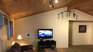 A living room with a leather sofa, a television on a stand, a lamp, and a wooden ceiling. - The Lake Geneva Lakehouse (Williams Bay)