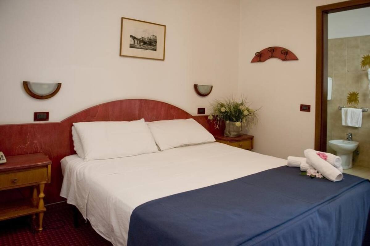A hotel room with a bed, bedside tables, a nightstand, a wall-mounted picture, a mirror, and a bathroom visible through an open door.