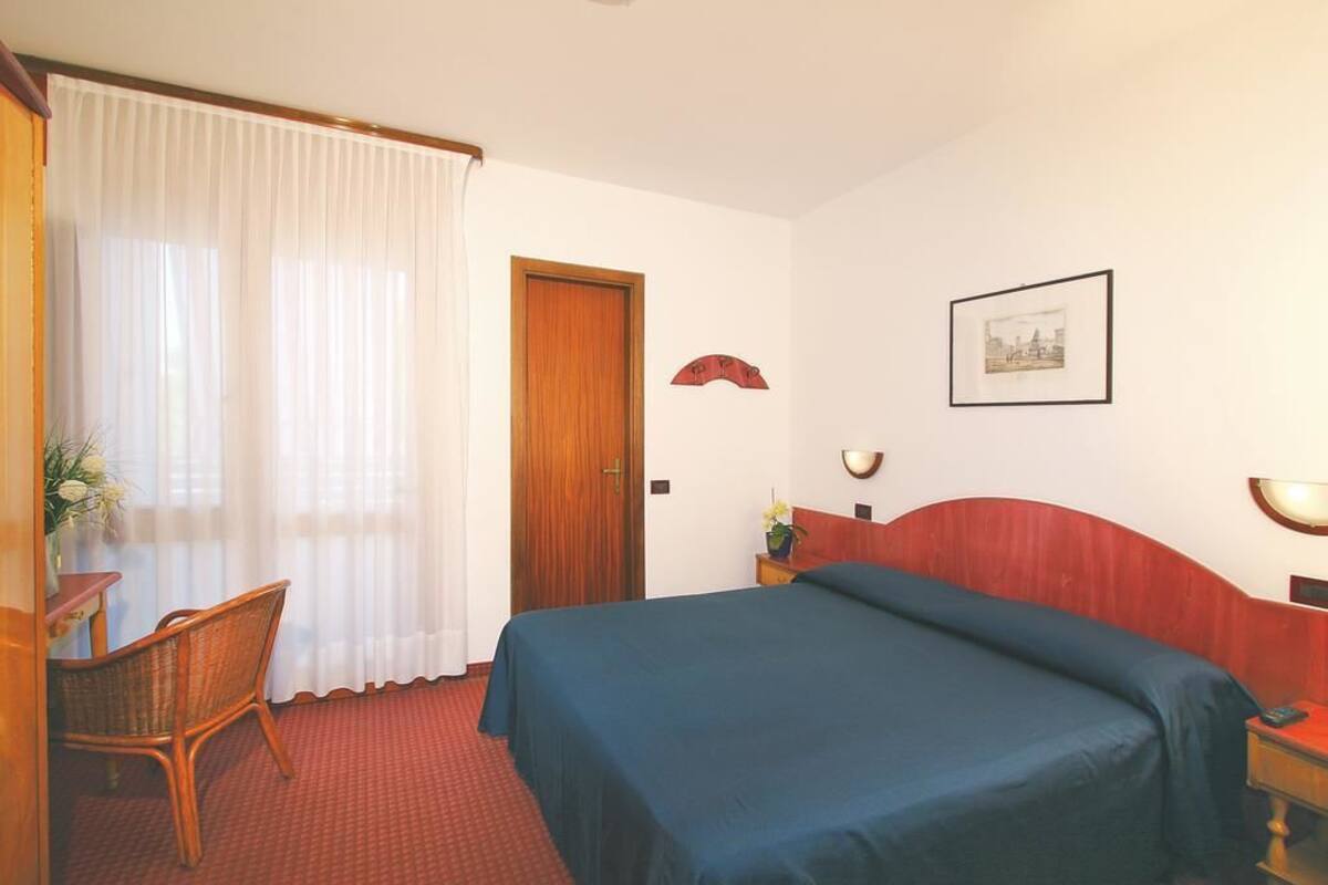 A hotel room with a bed, a chair, a desk, a door, and a wall-mounted picture.