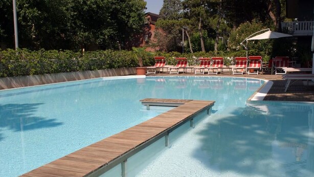 A clear swimming pool with a wooden deck and red and white lounge chairs. - Hotel Friuli (Lignano Sabbiadoro)