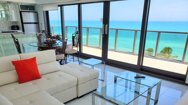 Un soggiorno moderno con un divano bianco, cuscini rossi e un tavolino da caffè in vetro. Dalle ampie finestre si vede un balcone con vista sull'oceano.
