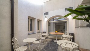 A small, enclosed patio area with white metal chairs and tables, a round table with a fruit bowl, and a view through an arched window. - Academia Resort (Bergamo)