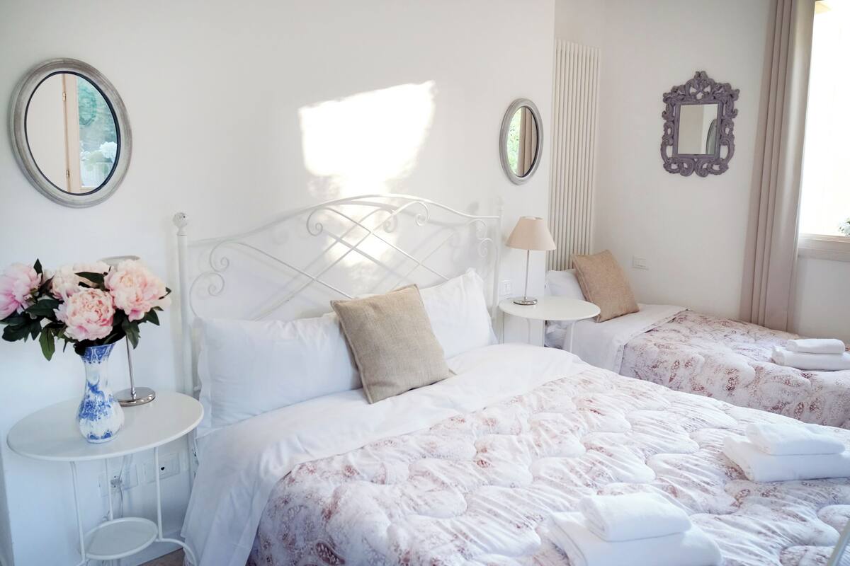 A bedroom with two beds, a bedside table with a lamp, a vase with flowers, and two wall mirrors.