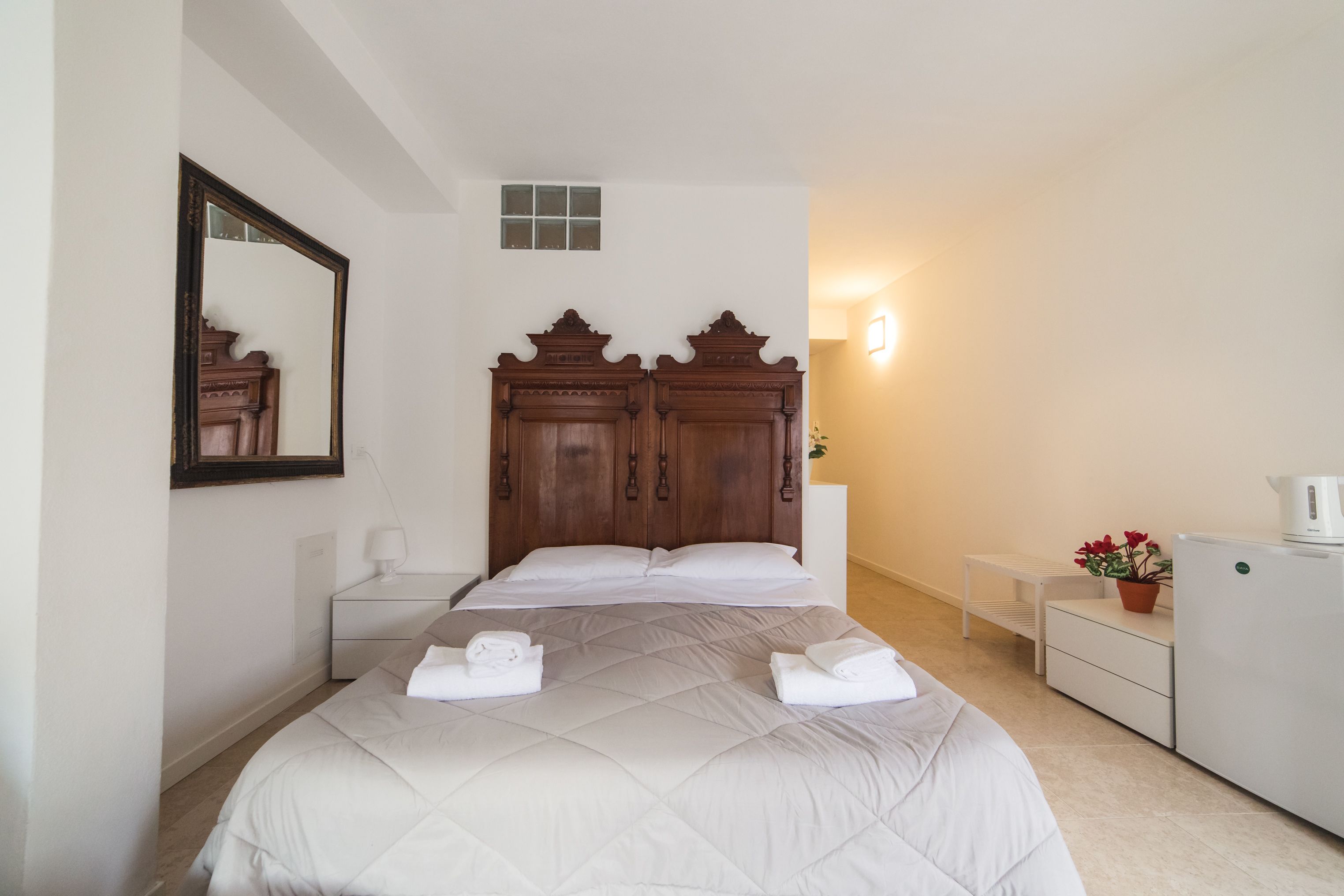 A hotel room with a large bed, a wooden headboard, a nightstand, a small table with a lamp, a potted plant, and a refrigerator.