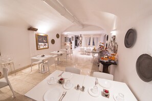 Un comedor con mesas y sillas blancas, una mesa de buffet con frutas y platos, y un espejo en la pared.
