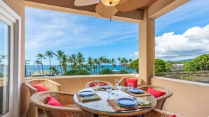A balcony with wicker furniture, a round table set for dining, and a view of the ocean and palm trees. - Aloha Condos, Beach Villas at Kahaluu, Villa 1-202, Beach View, AC (Kailua Kona)