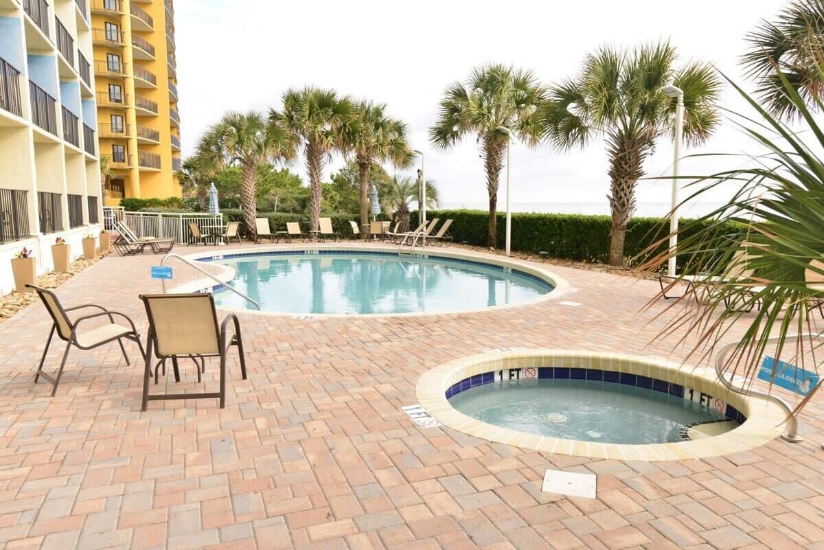 A pool area with lounge chairs and a hot tub, surrounded by palm trees and a building.