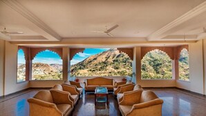 A room with a view of a mountainous landscape through arched windows, a ceiling fan, and a central seating area with sofas and a coffee table. - Kumbhalgarh Fort Resort (Kumbhalgarh)