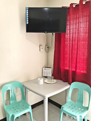 A small room with a TV mounted on the wall, a table with two chairs, a roll of toilet paper, and a menu board.