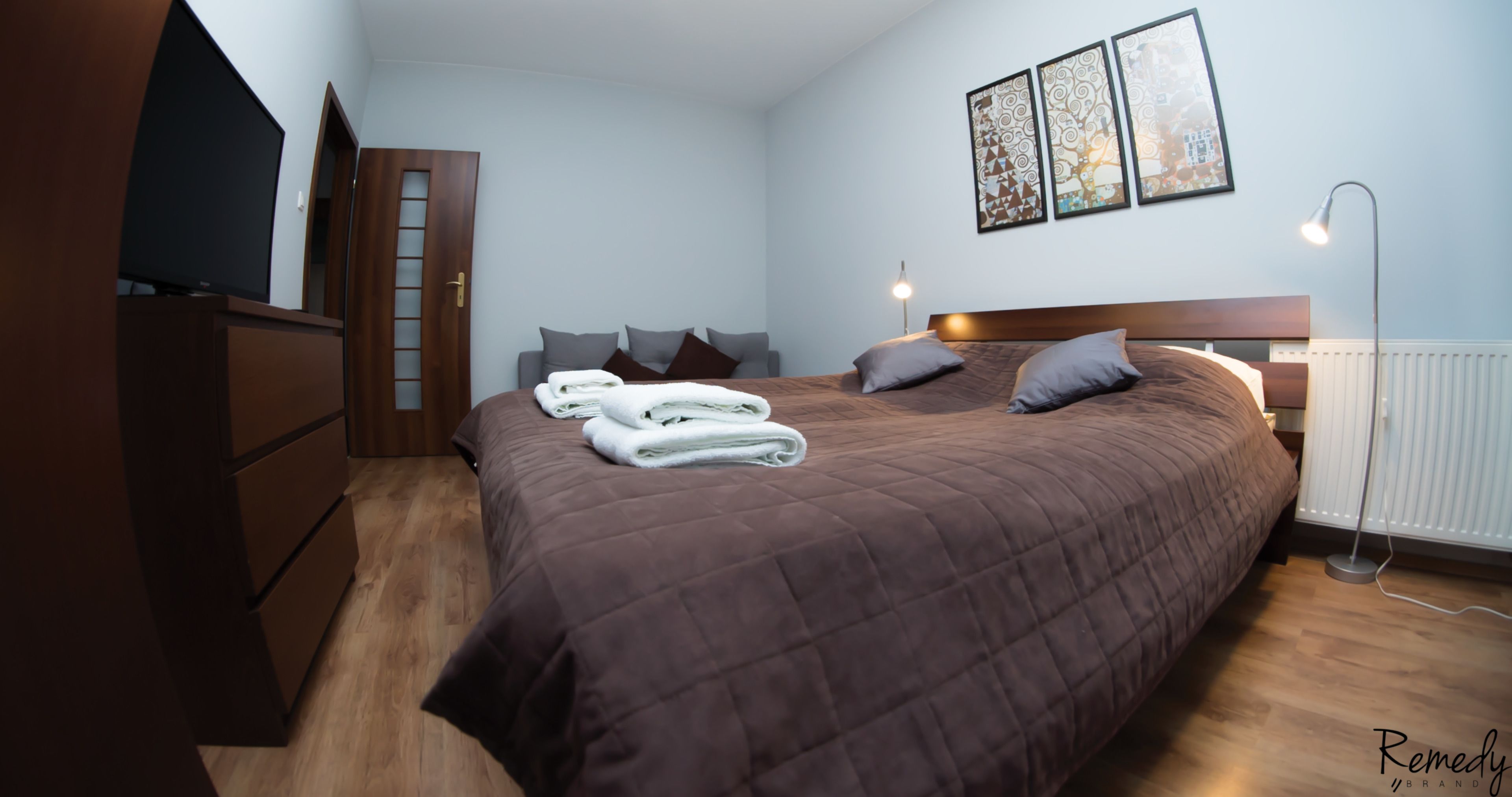 A neatly made bed with a brown quilt, a wooden dresser, a flat-screen TV, and a floor lamp.