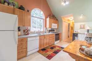 Una cocina con armarios de madera, un refrigerador blanco, un microondas y una estufa. Hay una ventana que proporciona luz natural y una alfombra en el suelo.