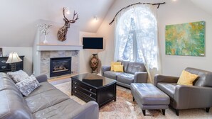 A modern living room with a fireplace, leather sofas, and a large window with a view of trees. - Sublime luxury  getaway riverfront downtown location great rates  (Estes Park)