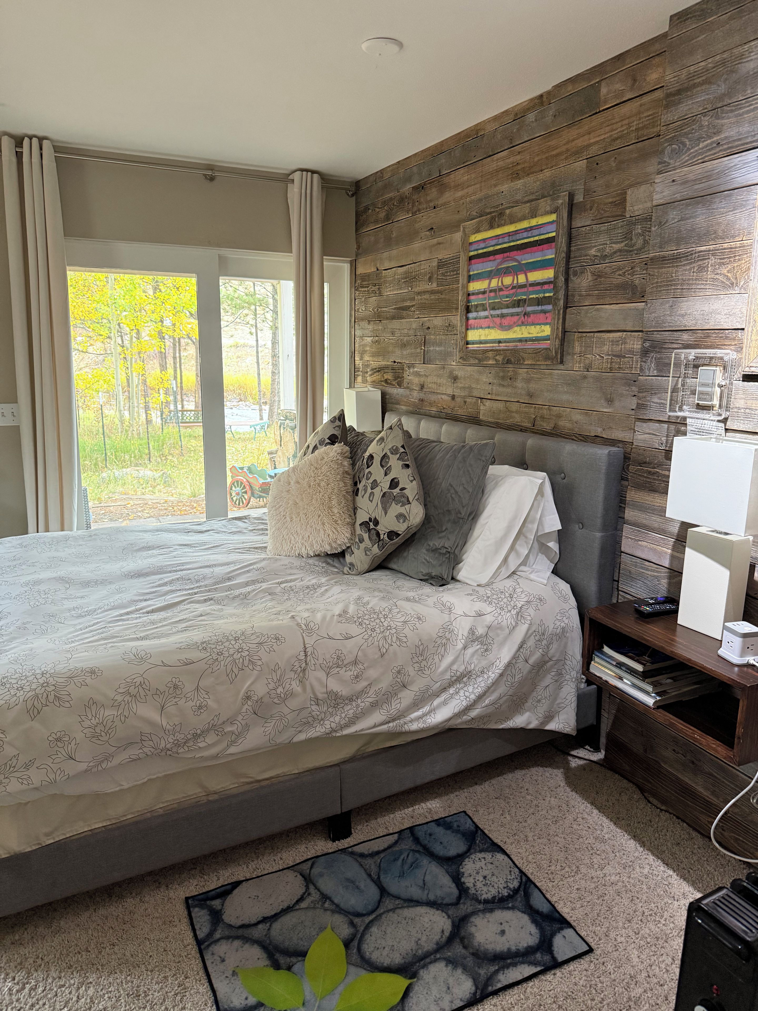 A bedroom with a bed, pillows, a nightstand, a lamp, a picture frame, and a view of the outdoors.