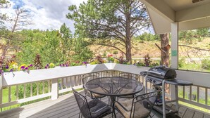 A covered outdoor patio with a round metal table and chairs, a grill, and a view of a river and trees. - Sublime luxury  getaway riverfront downtown location great rates  (Estes Park)