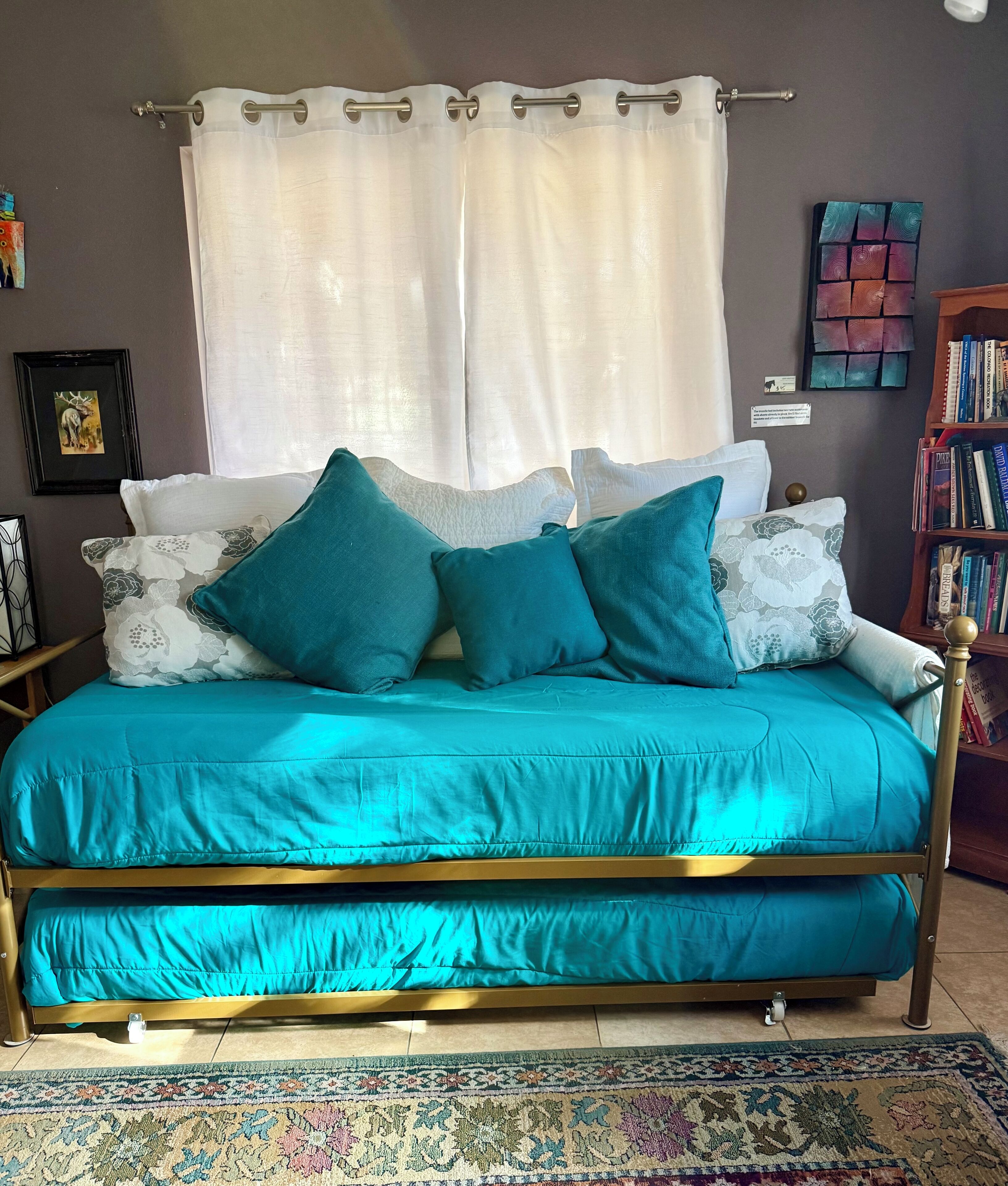 A bed with a teal bedspread and gold frame, a white curtain, and a patterned rug.