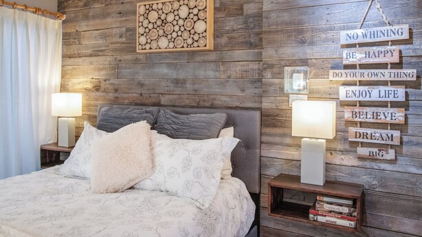 A bedroom with a wooden wall, a bed with floral bedding, two bedside lamps, and motivational wall decor. - Sublime luxury  getaway riverfront downtown location great rates  (Estes Park)