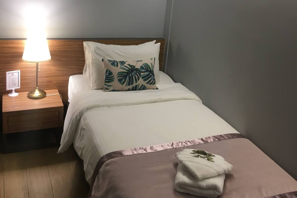 A hotel room with a bed, a nightstand with a lamp, a wall-mounted TV, and a folded towel on the bed.