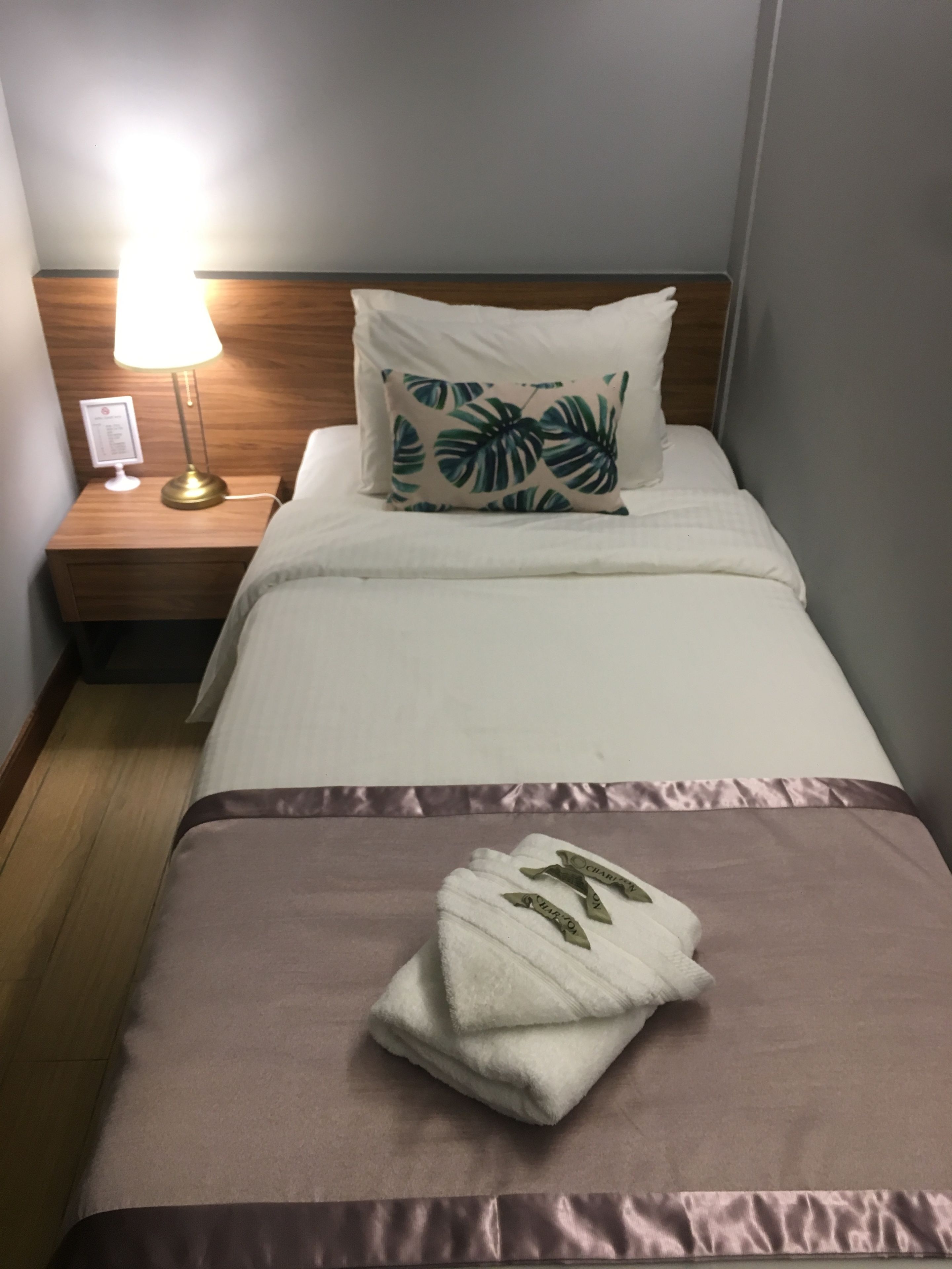 Un lit bien fait, recouvert d’une couverture marron, avec un oreiller blanc orné d’un motif de feuilles tropicales et une serviette blanche pliée, agrémentée d’un ruban vert, dans une chambre d’hôtel.