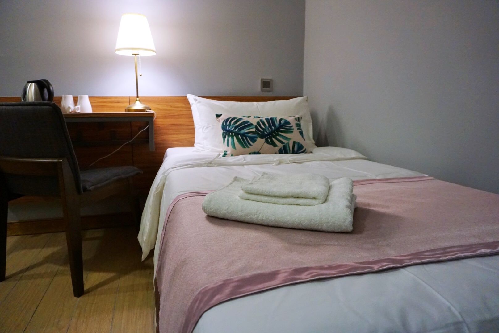 Quarto de hotel com uma cama, uma escrivaninha, uma cadeira, um abajur e uma cafeteira.