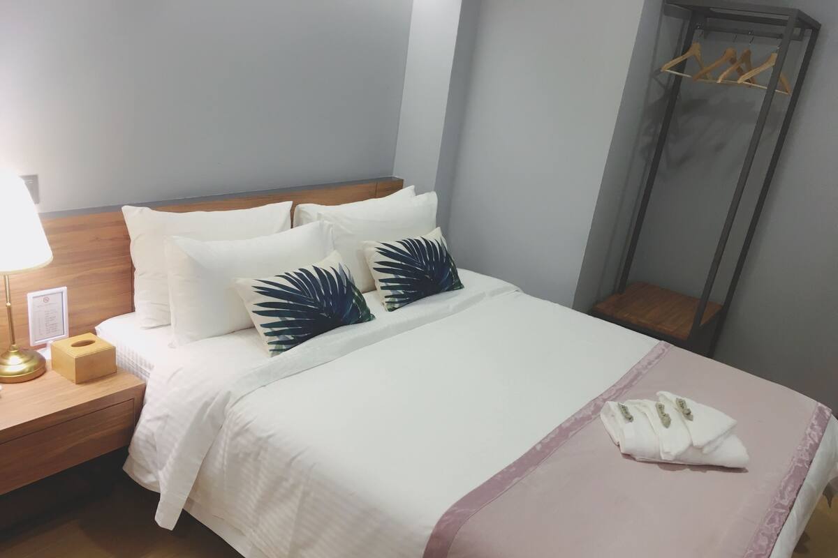 A neatly made bed with white bedding and two decorative pillows, a bedside table with a lamp and a tissue box, and a wooden nightstand.