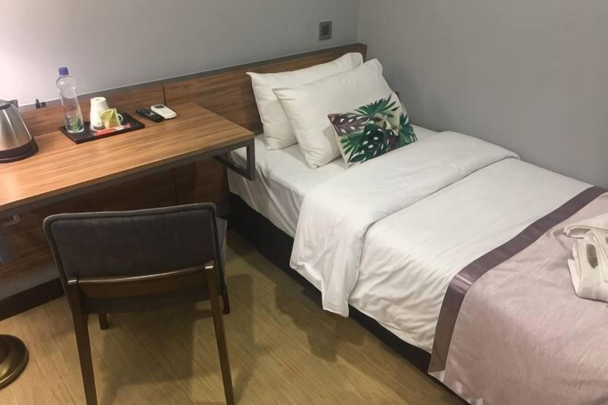 A hotel room with a wooden desk, a chair, a bed with a white and pink bedspread, a pillow with a tropical design, a water bottle, a kettle, and a mobile phone on the desk.