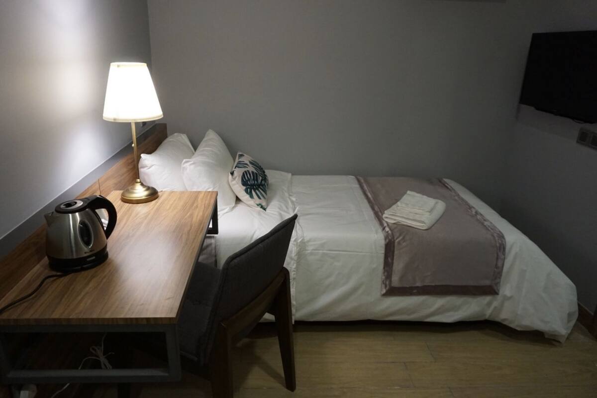 A small hotel room with a single bed, a bedside table with a kettle, a lamp, and a TV mounted on the wall.