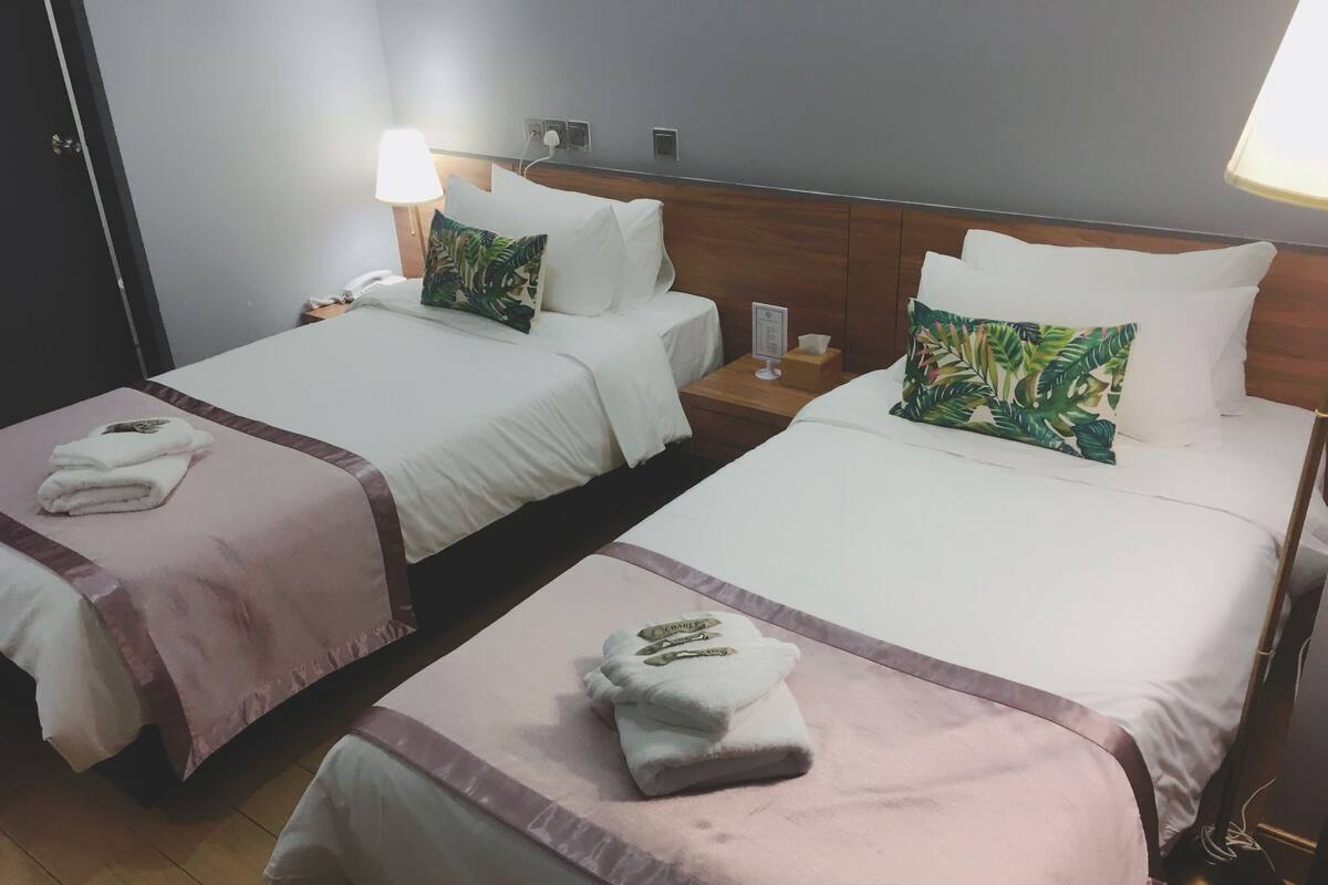 A hotel room with two beds, each with a white bedspread, pillows, and a small bedside table with a lamp and a phone.