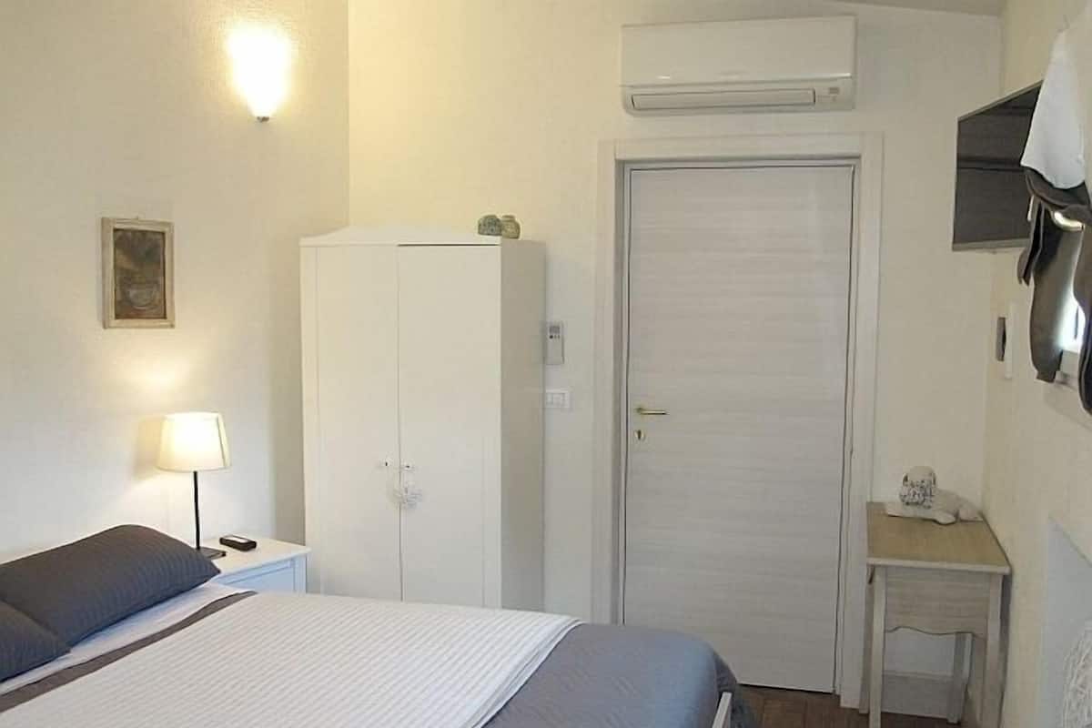 A small, neatly arranged bedroom with a bed, bedside table, lamp, wardrobe, and a small desk with a chair.