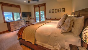 Un dormitorio con una cama grande, cabecera de madera, ventilador de techo, cómoda, televisor y vistas al exterior a través de amplias ventanas.