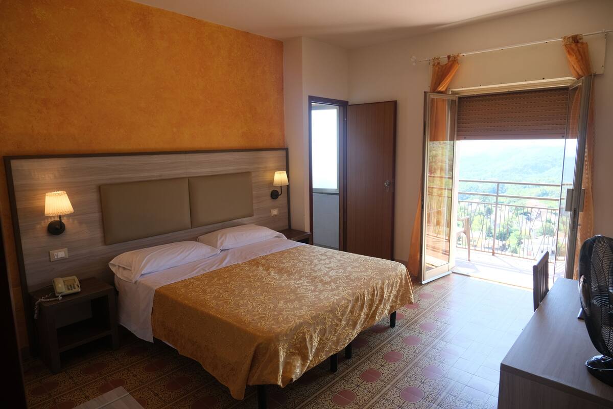 A hotel room with a bed, bedside lamp, phone, and a balcony with a view.